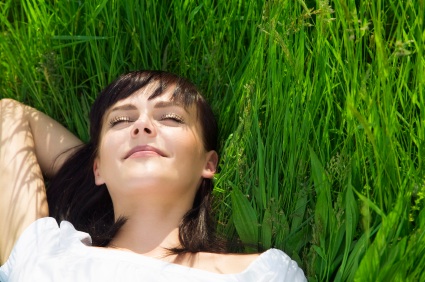 Young woman lying on grass with eyes closed, relaxing outdoors in sunlight.