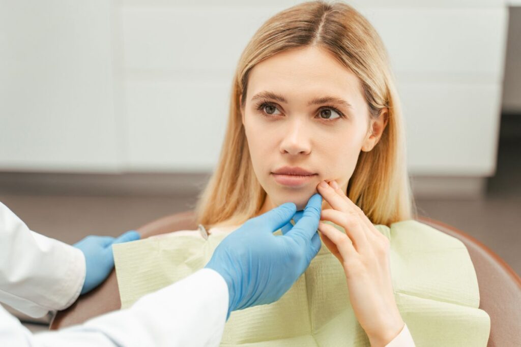 Female patient with reddish-brown hair receiving oral examination from healthcare provider wearing blue gloves in clinical setting.
