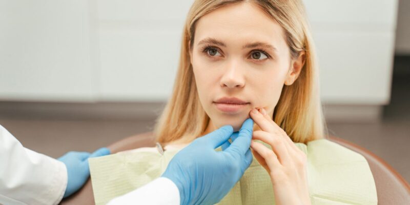 Female patient with reddish-brown hair receiving oral examination from healthcare provider wearing blue gloves in clinical setting.
