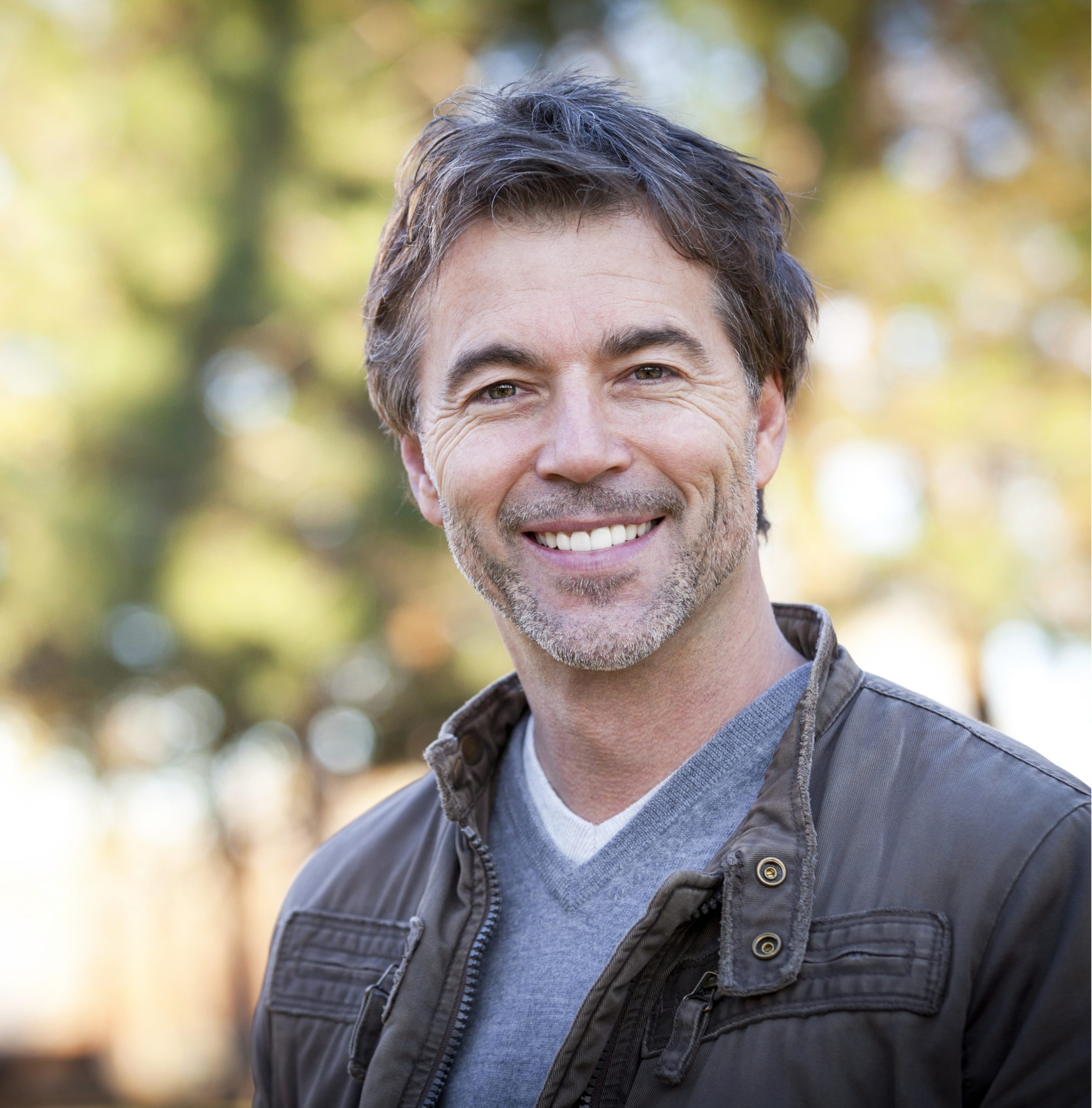 Man smiling at camera outdoors wearing dark jacket and blue shirt, blurred trees background.