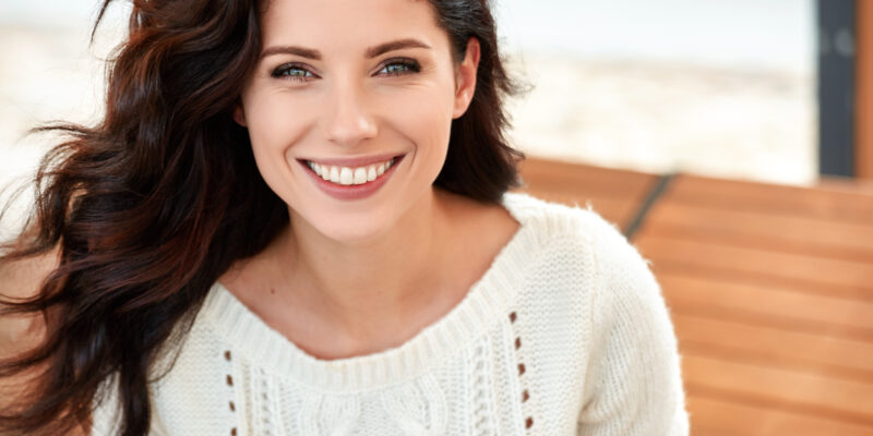 Smiling woman with dark wavy hair wearing cream sweater in bright indoor setting.