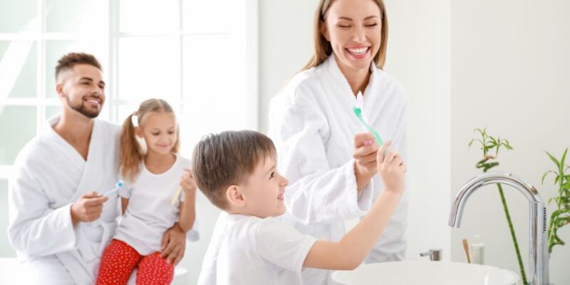 Family brushing teeth together in bright bathroom with mother helping young boy at sink.