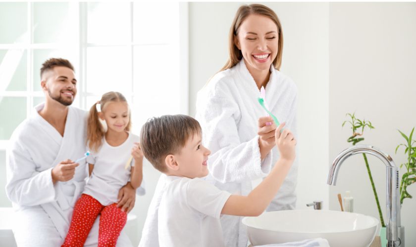 Family brushing teeth together in bright bathroom with mother helping young boy at sink.